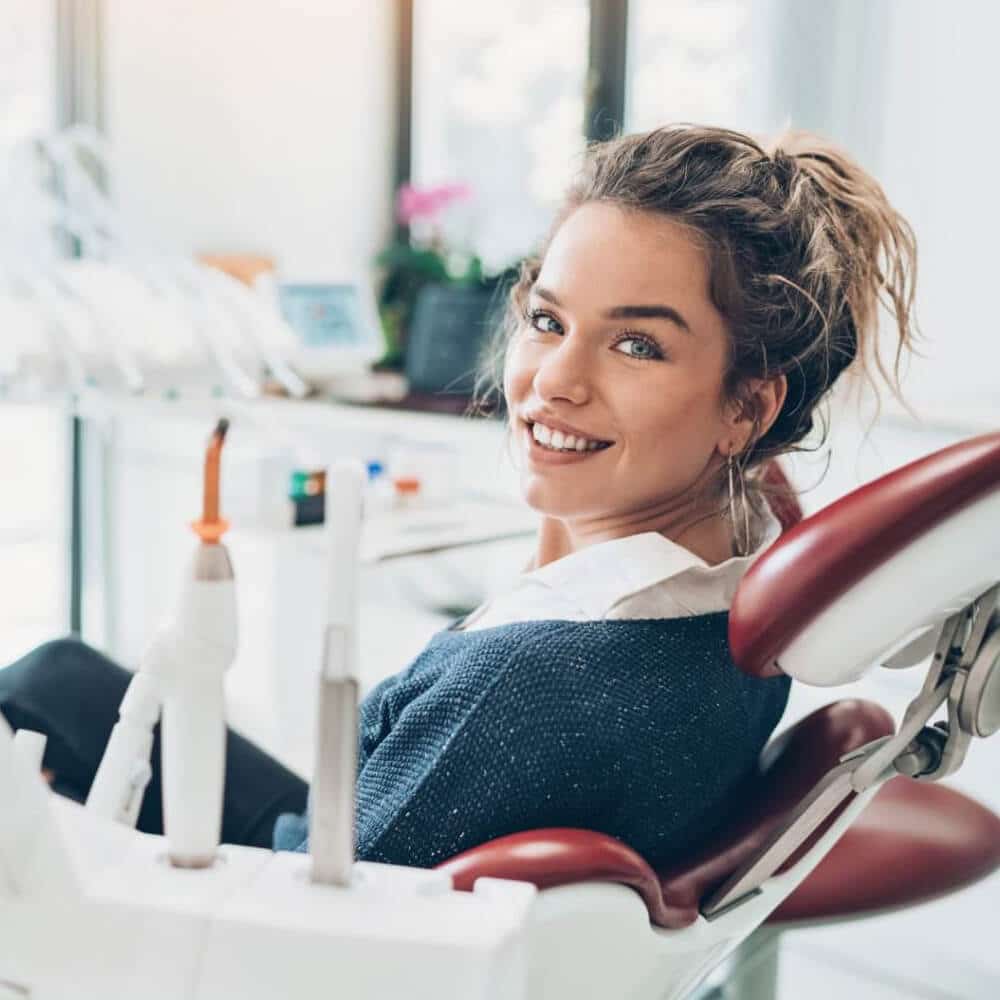 Young woman smiling in dental chair at modern dental office with equipment visible in background.