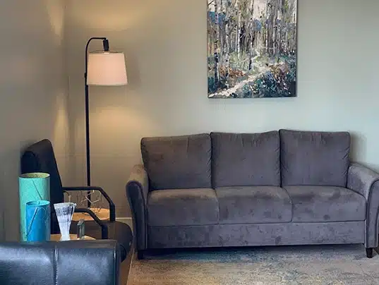 Modern dental office waiting room with gray sofa, floor lamp, and landscape artwork on wall.