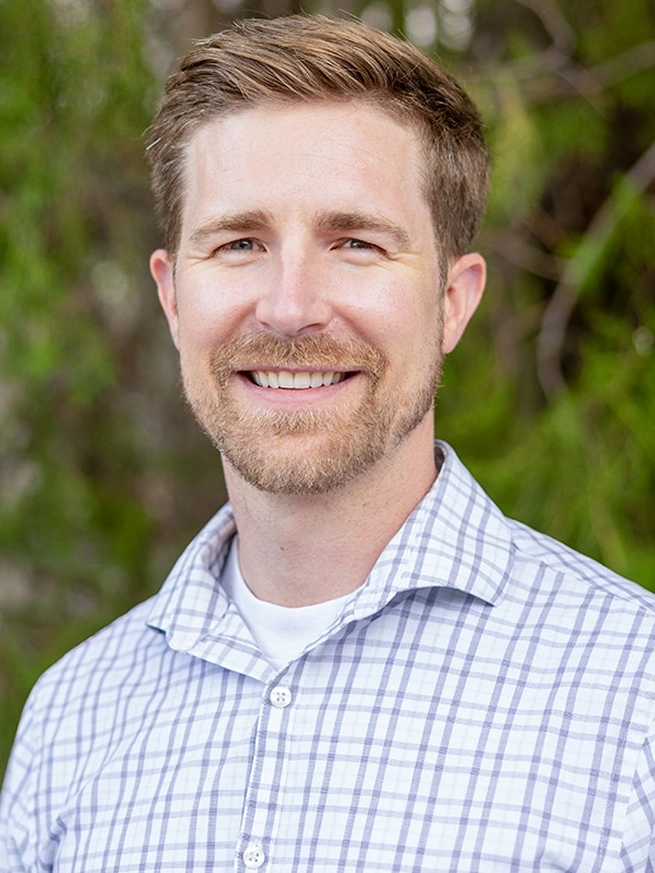 Professional headshot of smiling male dentist wearing checkered shirt against green foliage background.